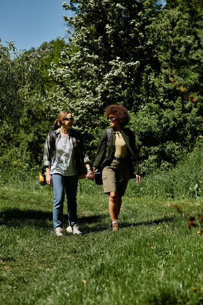 İki genç kadın, biri beyaz biri siyah, yemyeşil bir ormanda el ele yürürler, birlikte yürüyüş yaparlar.. - Fotoğraf, Görsel