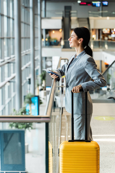 Mujer de negocios con valise mirando en ventana panorámica mientras espera en el embarque de vuelo de vacaciones. Chica asiática en traje de negocios en la terminal del aeropuerto con maleta amarilla mirando en la ventana - Foto, imagen