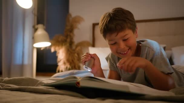 Счастливый милый сосредоточенный кавказский мальчик ребенок, маленький школьник, читающий книжную литературу, сын лежит кровать, учится дома, используя проведение электронного факела вечером перед сном освещенная спальня внутри - Кадры, видео