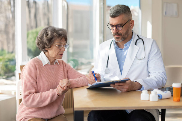 Άνδρας γιατρός με στολή με στηθοσκόπιο κρατώντας έγγραφα, συμβουλεύεται ηλικιωμένη γυναίκα σχετικά με το αποτέλεσμα του ελέγχου, ώριμος ασθενής ακούει γιατρό εξηγώντας συνταγή - Φωτογραφία, εικόνα
