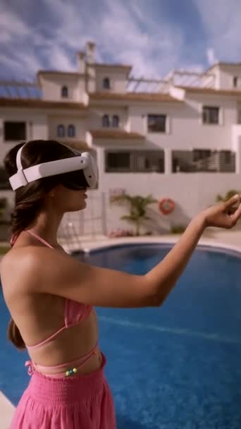 Mujer con un casco VR y un traje de baño rosa, experimentando la realidad virtual junto a una piscina en un resort soleado. - Metraje, vídeo