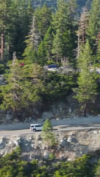 Вертикальный снимок: захватывающий вид на автомобили, проносящиеся по живописной горной дороге возле Лейк-Плэсид, Калифорния, в окружении пышных сосен и скалистых образований. - Кадры, видео