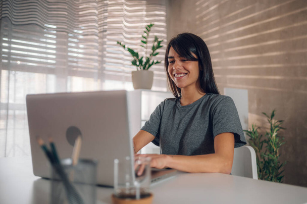 Retrato de uma mulher de negócios feliz digitando um e-mail em seu laptop enquanto se senta em sua mesa em um escritório em casa adorável e ensolarado. Concentre-se em seu rosto satisfeito e feliz. Conceito de espaço de trabalho agradável. - Foto, Imagem