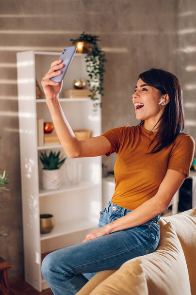 Uśmiechnięta kobieta używająca smartfona do robienia selfie podczas relaksu w domu. Piękna młoda dama robi zdjęcia uśmiechając się i patrząc w kamerę telefonu komórkowego. Przestrzeń kopiowania. - Zdjęcie, obraz