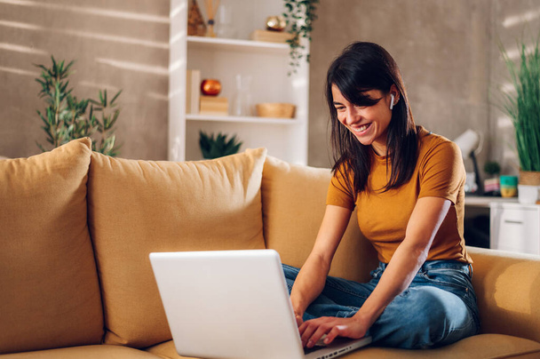 Portret kobiety korzystającej z laptopa i sprawdzającej wiadomości e-mail lub wiadomości online, siedzącej na kanapie w domu. Poszukiwanie przyjaciół w internetowych sieciach społecznościowych lub praca na komputerze. - Zdjęcie, obraz