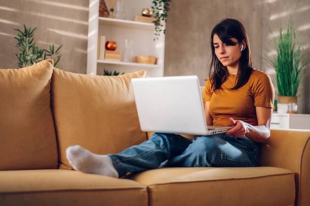 Молодая женщина работает на ноутбуке и улыбается, сидя дома на диване. Симпатичная девушка сидит дома на диване и управляет своим бизнесом. - Фото, изображение