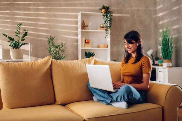 Retrato de uma mulher usando airpods laptop e verificar e-mail ou notícias on-line enquanto sentado no sofá em casa. Buscando amigos na internet redes sociais ou trabalhando no computador. Espaço de cópia. - Foto, Imagem