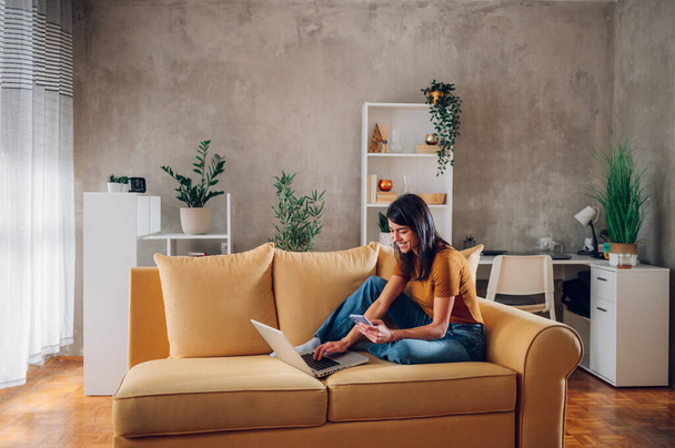 Schöne junge Frau, die am Laptop arbeitet und ein Smartphone benutzt, während sie zu Hause auf der Couch sitzt. Nettes Mädchen sitzt zu Hause auf dem Sofa und führt ihr Geschäft. - Foto, Bild