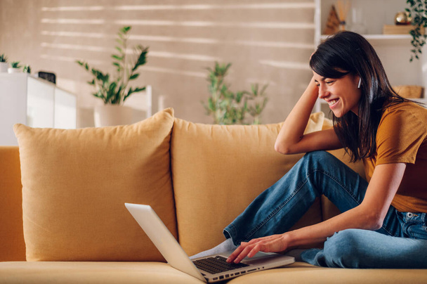 Sivukuva hymyilevästä valkoihoisesta naisesta, joka käyttää kannettavaa tietokonetta istuessaan sohvalla kotona. Onnellinen naisten ostoksia tai chattailuun verkossa sosiaalisessa verkostossa, katsomassa videota tai elokuva. Kopioi tila. - Valokuva, kuva