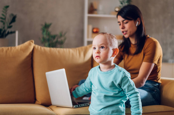 Szerető fiatal anya élvezi a hétvégét a kisbabájával, miközben játszik vele és laptopot használ. Gondoskodó, boldog anya jól érzi magát a kisfiús gyerekkel. Koncentrálj a portréra. - Fotó, kép