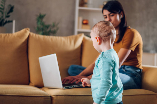 Memnuniyetle anne ya da bebek bakıcısı gülümseyen ve kucak kucak açan çocuk evde kanepede dinlenirken ve dizüstü bilgisayar kullanırken. Mutlu aile hayatı. Küçük bir çocuğun arka planına odaklan.. - Fotoğraf, Görsel