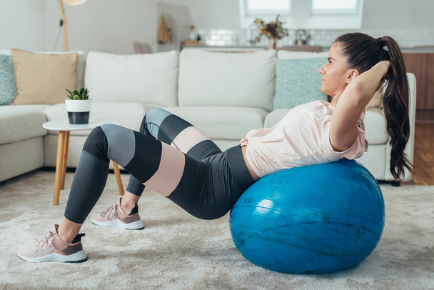Junge attraktive Frau knirscht beim Training zu Hause auf einem Fitnessball - Foto, Bild