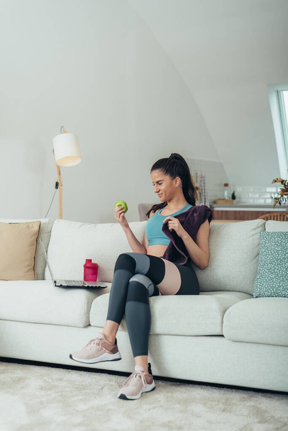 Mladá žena sedí na pohovce po tréninku doma při používání notebooku a jíst jablko - Fotografie, Obrázek