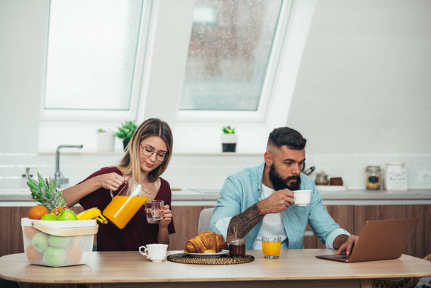Jeune couple attrayant prenant le petit déjeuner dans la cuisine à la maison tout en utilisant un ordinateur portable - Photo, image