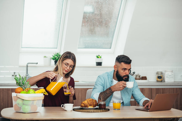 Νεαρό ζευγάρι παίρνει πρωινό στο σπίτι ενώ χρησιμοποιεί ένα φορητό υπολογιστή και πίνοντας καφέ και χυμό πορτοκαλιού - Φωτογραφία, εικόνα