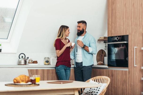 Young beautiful couple drinking coffee in the kitchen and spending time together in the morning - Photo, Image