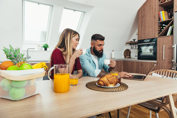 Съемка молодой счастливой пары, наслаждающейся завтраком дома, используя ноутбук и пить кофе - Фото, изображение