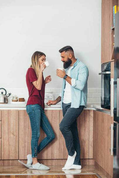 コーヒーを飲みながら自宅のキッチンでリラックスしている愛情のある若いカップルのショット - 写真・画像