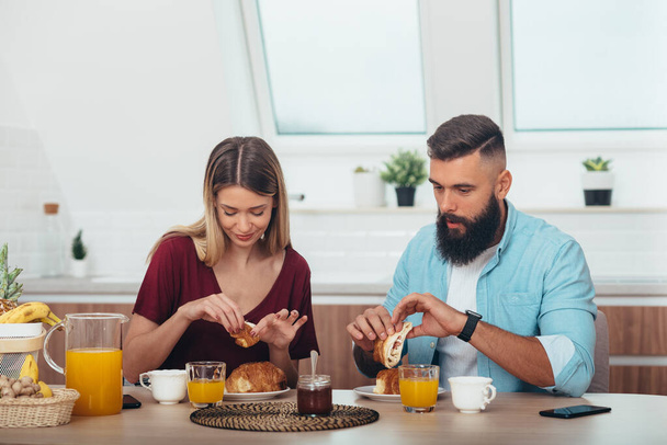 Casal afetuoso ter croissants para o café da manhã na cozinha em casa enquanto bebe café e suco de laranja - Foto, Imagem