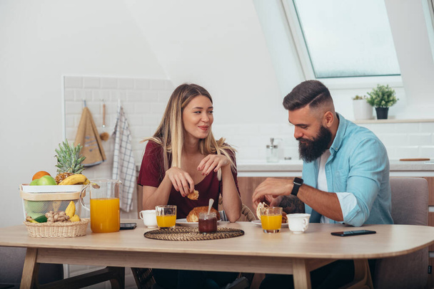 Aantrekkelijk koppel ontbijten in de keuken thuis terwijl ze croissants eten en sinaasappelsap drinken - Foto, afbeelding