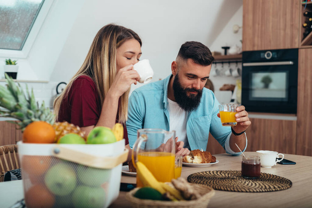 Foto de una joven pareja desayunando en la cocina de su casa mientras come croissants y bebe café y zumo de naranja - Foto, imagen