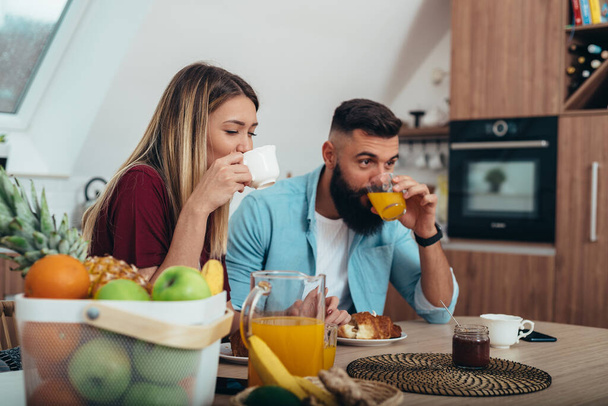 Aufnahme eines jungen Paares, das zu Hause in der Küche frühstückt, während es Kaffee und Orangensaft trinkt - Foto, Bild