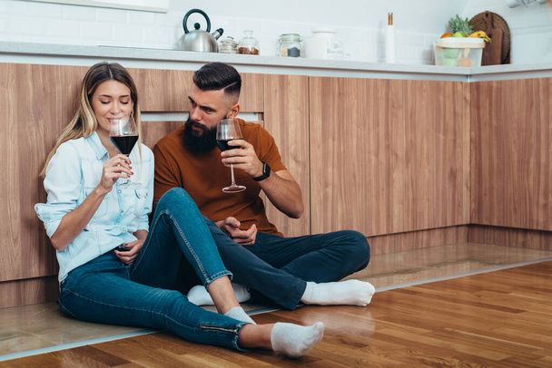 Belo casal bebendo vinho na cozinha em casa, enquanto sentado no chão e tendo um momento romântico apenas para eles - Foto, Imagem