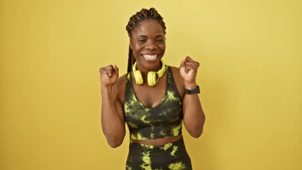 Mujer afroamericana triunfante en ropa deportiva, brazos levantados en celebración de la victoria, exudando confianza y alegría sobre un fondo amarillo aislado - Imágenes, Vídeo