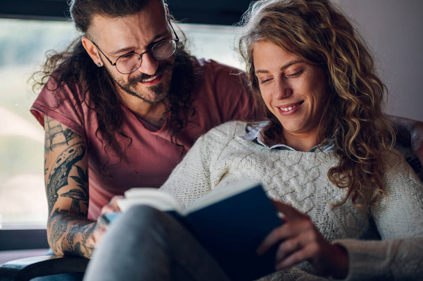Close shot of a young couple at home. Man is embracing his girlfriend while she is reading a book and sitting on chair. Two people relaxing together at home and spending free time together. - Photo, Image