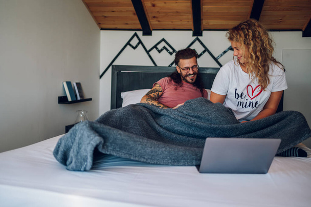 愛情深い若いカップルは,朝,毛布で覆われたベッドルームに座っています. ベッドでラップトップを使用して,朝のニュースやインターネットを閲覧するカップル. あなたの愛する人と一日を始める. - 写真・画像