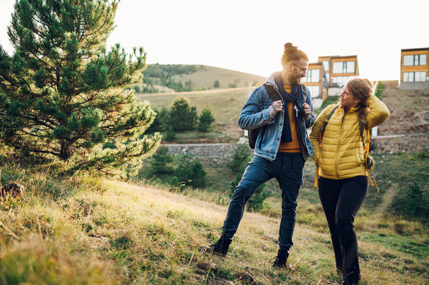 Ρομαντικό ζευγάρι τουριστών ντυμένοι με ζεστά ρούχα περπατώντας σε ένα καταπράσινο βουνό κορυφογραμμή κατά τη διάρκεια ενός ηλιοβασιλέματος με ένα αστικό σπίτι στο παρασκήνιο. Νέοι που περπατούν στη φύση. - Φωτογραφία, εικόνα