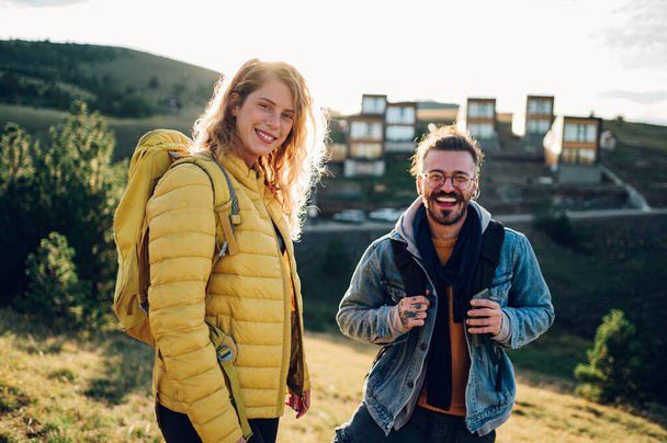 ハイキングギアの旅行カップルは,カメラを見ながら日没の山を歩いています. 笑顔の配偶者が丘の下を歩き,日没の間に自然を楽しんでいる. - 写真・画像