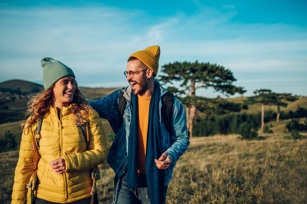 暖かい服を着た観光客の笑顔のカップルの肖像画は,日没の間に草の多い山の尾根を歩いています. 自然の中でハイキングをする若者たち. 男は自然の中で楽しんでいる間に女性を抱きしめる. - 写真・画像