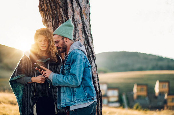 Portrét bělošského páru, který odpočívá pod stromem a používá spolu smartphone. Manželé pěší turistiku v horách a užívají si dobrodružství na dovolené. Aktivní životní styl. Kopírovat prostor. - Fotografie, Obrázek