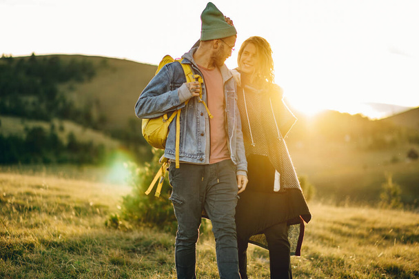 Happy young couple having fun on their hiking trip while walking together on a mountain trail with a sun flare behind them during sunset. Caucasian hiker couple enjoying themselves on a vacation. - Photo, Image