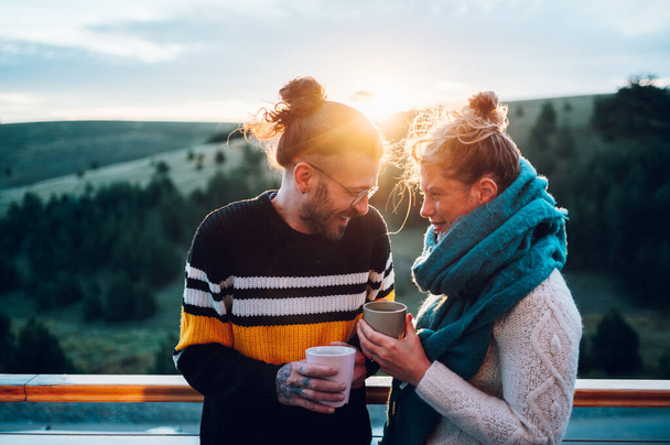 Happy couple enjoying morning coffee and looking at each other while standing on a balcony with an beautiful nature in the background and with a warm light at sunrise. Cold weather. - Photo, Image