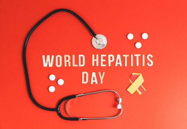 Un estetoscopio, pastillas blancas, una cinta de color amarillo-rojo y letras de madera clara con la frase forrada Día Mundial de la Hepatitis se encuentran en el centro sobre un fondo rojo, plano de cerca. - Foto, imagen