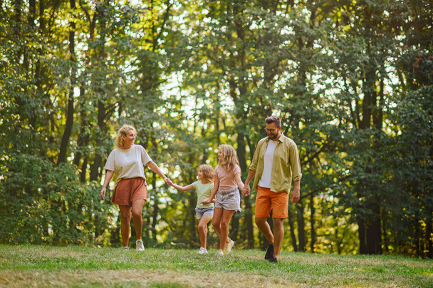 子供たちと手を握り,晴れた夏の日に自然の中で質の高い時間を過ごす幸せな両親. 晴れた日にカジュアルな散歩をしながら,陽気な家族の絆と自然を探検する. - 写真・画像