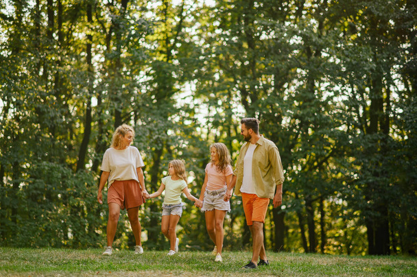 Full length of a happy family of four holding hands and walking on a meadow near forest in nature on sunny day. Cheerful family is spending weekend in nature together. Family is strolling in nature. - Photo, Image
