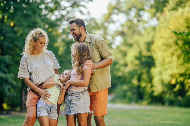A young family is standing together at the meadow near forest and hugging. Portrait of happy parents spending day in nature with their daughters. Affectionate family spending day outdoors on sunny day - Photo, Image