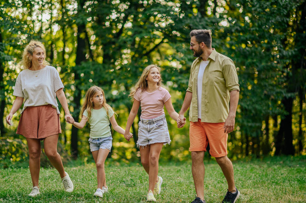 Portret kochającej rodziny spacerującej w przyrodzie i nawiązującej więź podczas weekendowego i słonecznego letniego dnia w przyrodzie. Rodzice trzymający się za ręce z córkami i spacerujący po łące w pobliżu lasu. - Zdjęcie, obraz