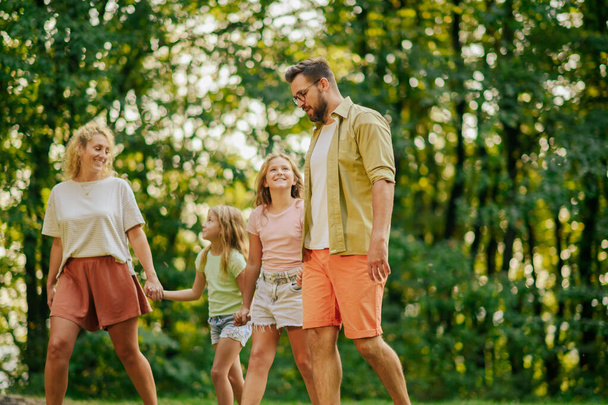 Una familia cariñosa de cuatro personas está cogida de la mano y dando un paseo por la naturaleza. Retrato de padres paseando en la naturaleza con sus hijas y hablando con ellas mientras exploran la naturaleza en el soleado día de verano. - Foto, imagen