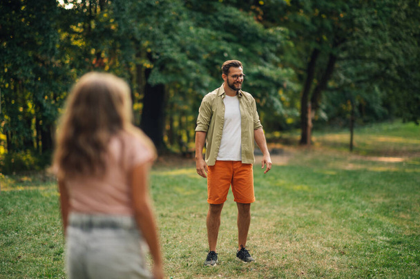 A young father is spending sunny summer day in nature with his beloved daughter in blurry foreground. Father having fun in nature while standing on a meadow near forest and laughing with child. - Photo, Image