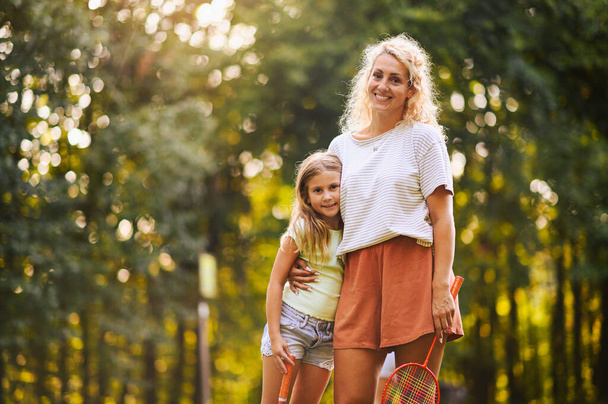 Portré egy boldog anya és lánya kezében tollaslabda ütők és állva a természetben, miközben ölelés mosolyogva a kamerába. Sportos, aktív család, akik napsütéses nyári napokat töltenek a természetben. Fénymásolási hely. - Fotó, kép