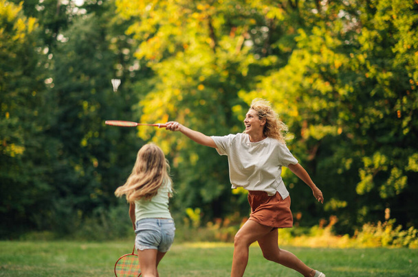 Egy sportos anya akcióban üti a tollaslabda ütővel, miközben tollaslabdázik a lányával a természetben, az erdő közelében, napsütéses nyári napon. Egy sportos család tollaslabdázik a természetben. - Fotó, kép
