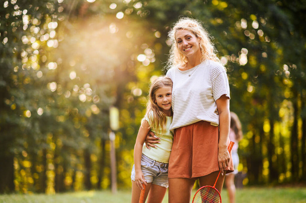 Portrait of a cheerful mother standing in nature with badminton racquet in her hand and hugging her daughter while smiling at the camera on sunny summer day. A happy mom and child on sports day. - Photo, Image