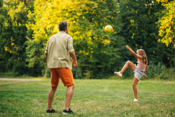 Ένας αθλητικός πατέρας και η κόρη παίζει μπάλα παιχνίδι στη φύση. Μια κόρη κλωτσάει μια μπάλα ενώ κάνει σπορ στο λιβάδι της φύσης. Μια αθλητική μέρα με τον πατέρα στη φύση. Ημέρα του πατέρα, πατρότητα. - Φωτογραφία, εικόνα