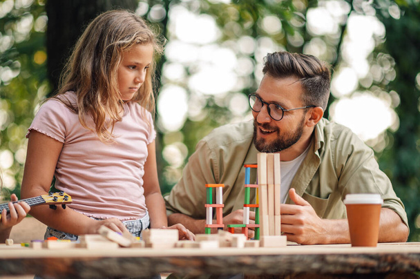 Ένας διασκεδαστικός μπαμπάς παίζει ένα διασκεδαστικό χτίσιμο ενός παιχνιδιού πύργου με την κόρη του, ενώ κάθεται στη φύση στο τραπέζι και έχει πικνίκ. Ένας πατέρας χαμογελάει και διασκεδάζει παίζοντας ένα παιχνίδι με την κόρη του στη φύση. - Φωτογραφία, εικόνα