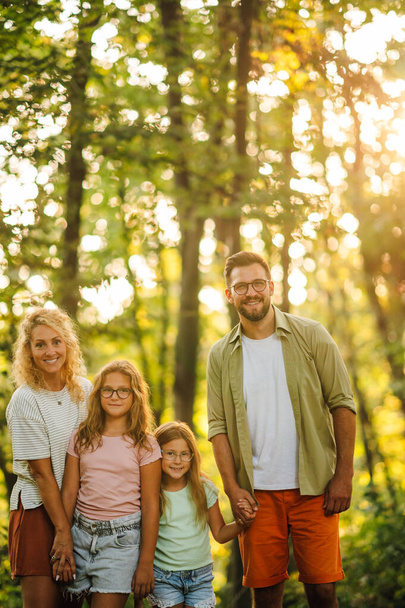 Egy boldog család portréja áll a természetben és mosolyog a kamerába. Boldog szülők fogják egymás kezét a gyerekeikkel, és napsütéses nyári napot töltenek a természetben. Erdőt felfedező család. - Fotó, kép
