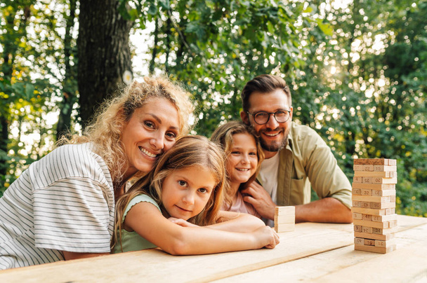 愛情深い家族が自然の中でピクニックのテーブルに座り,カメラで笑顔で抱きしめています. 愛する両親が娘を抱きしめる. ジェガゲームで自然にポーズする家族. 家族の価値観 - 写真・画像